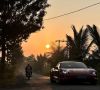 Roter Porsche in Frontaufnahme neben Motorrad auf Straße im Sonnenuntergang, zu beiden Seiten Bäume, Sträucher und Palmen